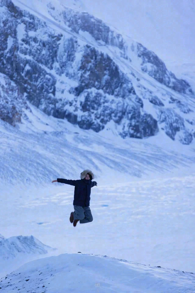 A person jumps with arms extended against a snow-covered mountain face.