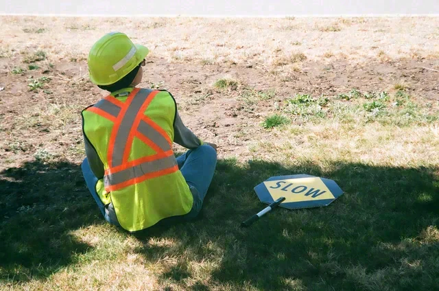 A person wearing a bright safety vest and green hard hat sits on grass beside a sign marked SLOW.
