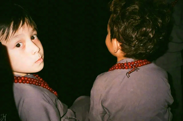 Two young people wear red beaded necklaces in a dark setting; one turns to look toward the camera.