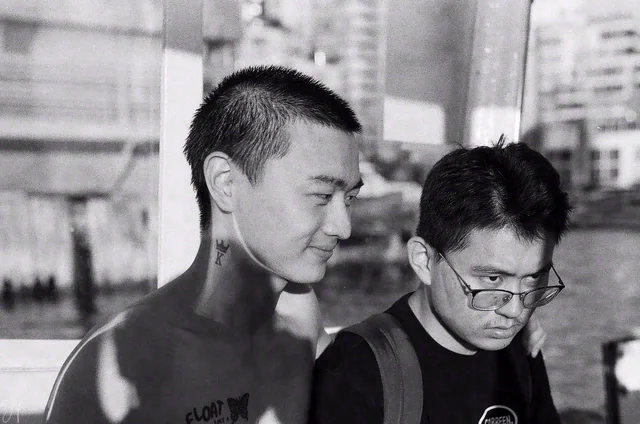 Black and white close-up of two people, one looking sideways and one wearing glasses and looking downward.