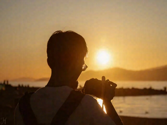 A backlit person holds a camera in front of their chest, with golden sunset light over the water behind them.