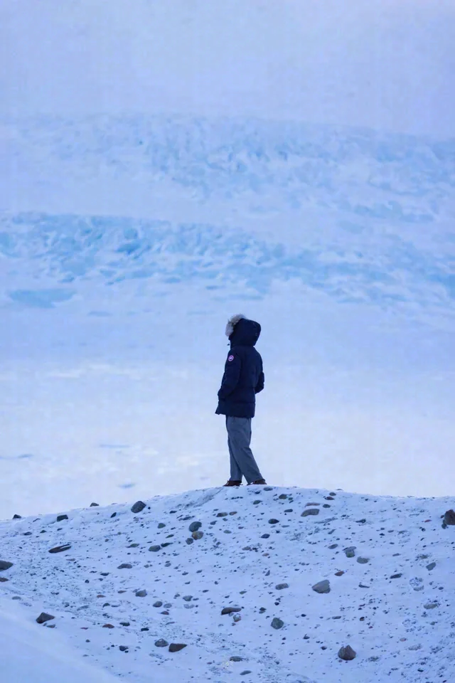 A person stands in profile on a snowy ridge, with pale blue snow-covered slopes behind them.