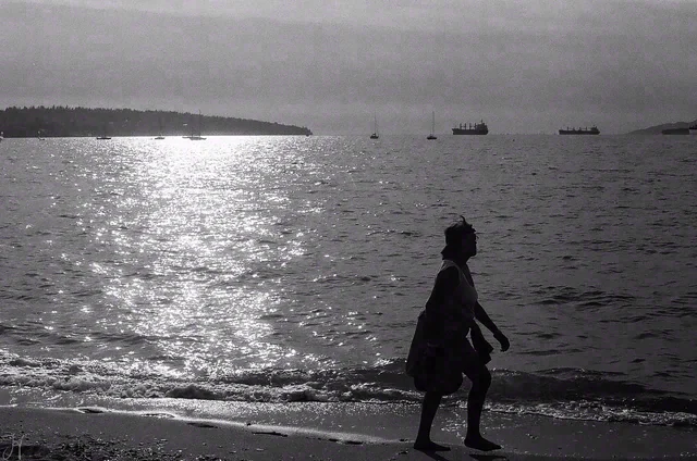 Black and white silhouette of a person walking beside water sparkling in the sun, with boats in the distance.