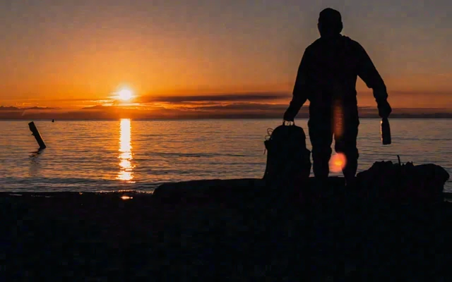 A silhouetted person stands at the shoreline beneath an orange sunset sky, with sunlight reflected across the water.