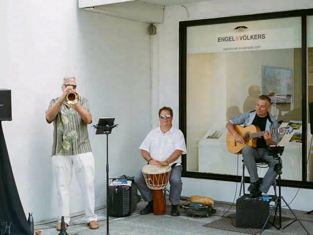 Three people play a trumpet, a hand drum and an acoustic guitar outside a building.